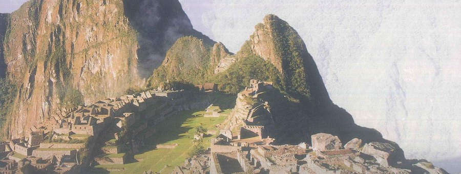 Stone ruins and green terraces on a narrow mountain ridge, with steep peaks and clouds behind them