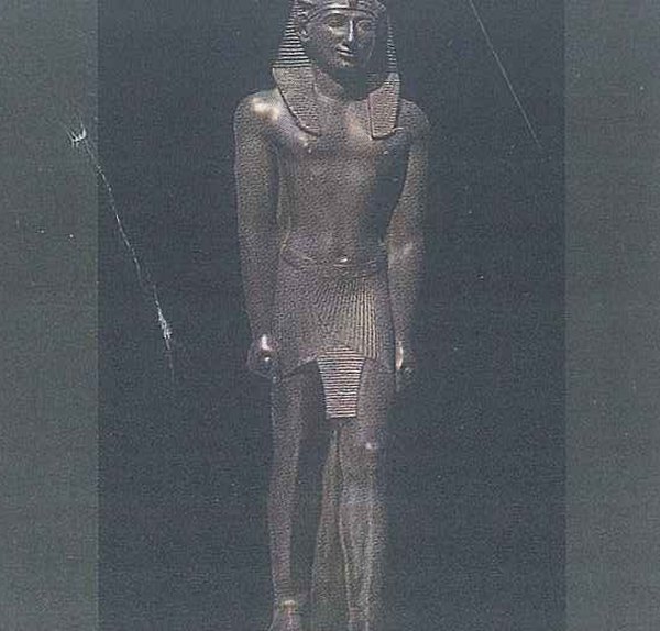 A dark statue of a standing man in a striped headdress and a short pleated skirt, arms by his sides