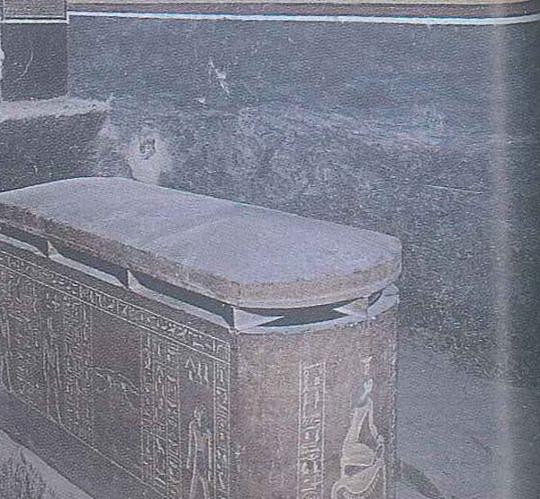 A large carved box with a heavy flat lid in a dark room, its sides covered in small figures and signs